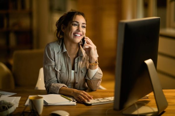 young-happy-freelancer-communicating-mobile-phone-while-surfing-net-desktop-pc-night-home